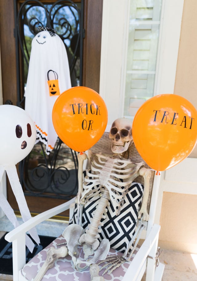 Halloween porch display with themed helium balloons, pumpkins and a seated skeleton arranged near the door