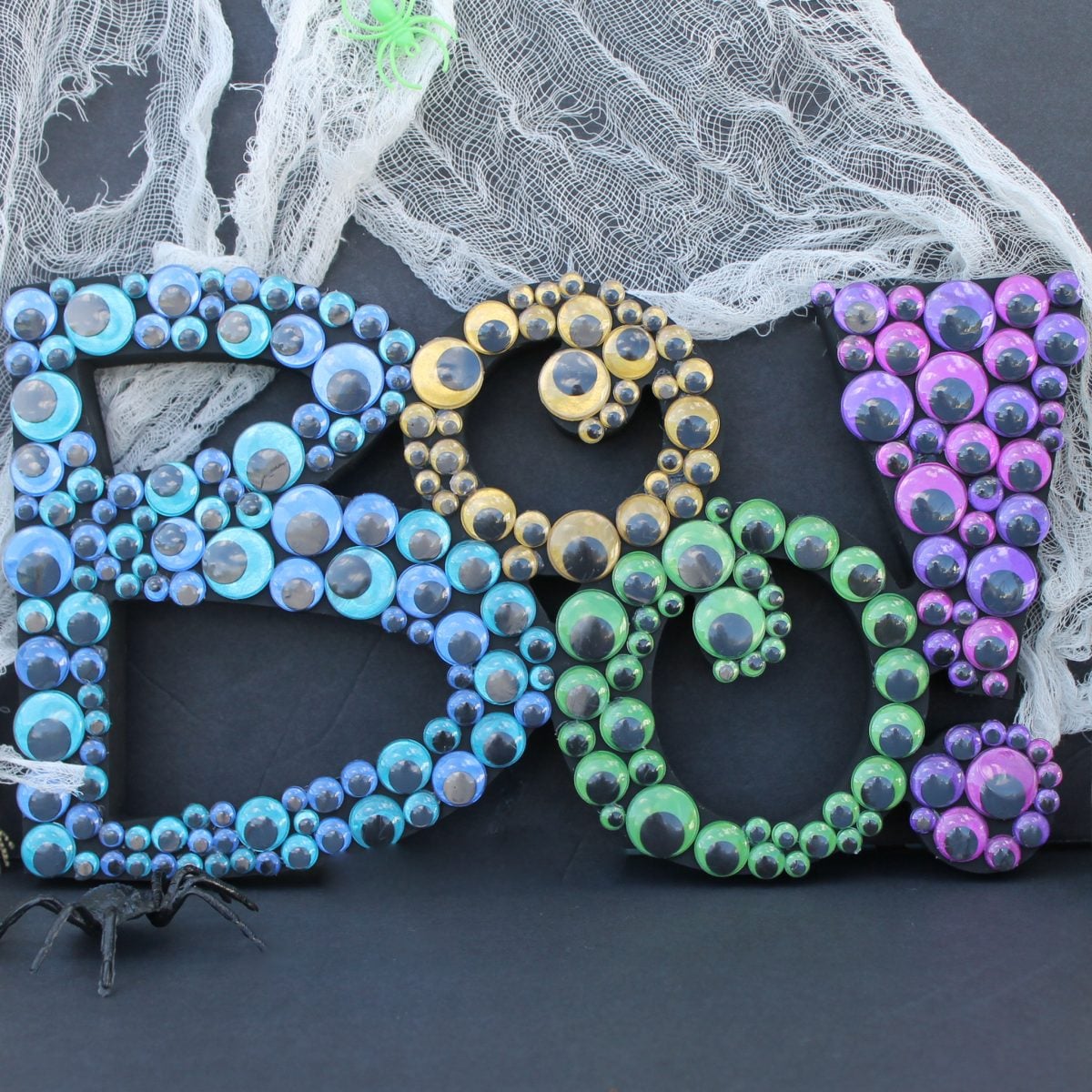 Black BOO sign covered with metallic blue gold green and purple googly eyes rests against dark fabric