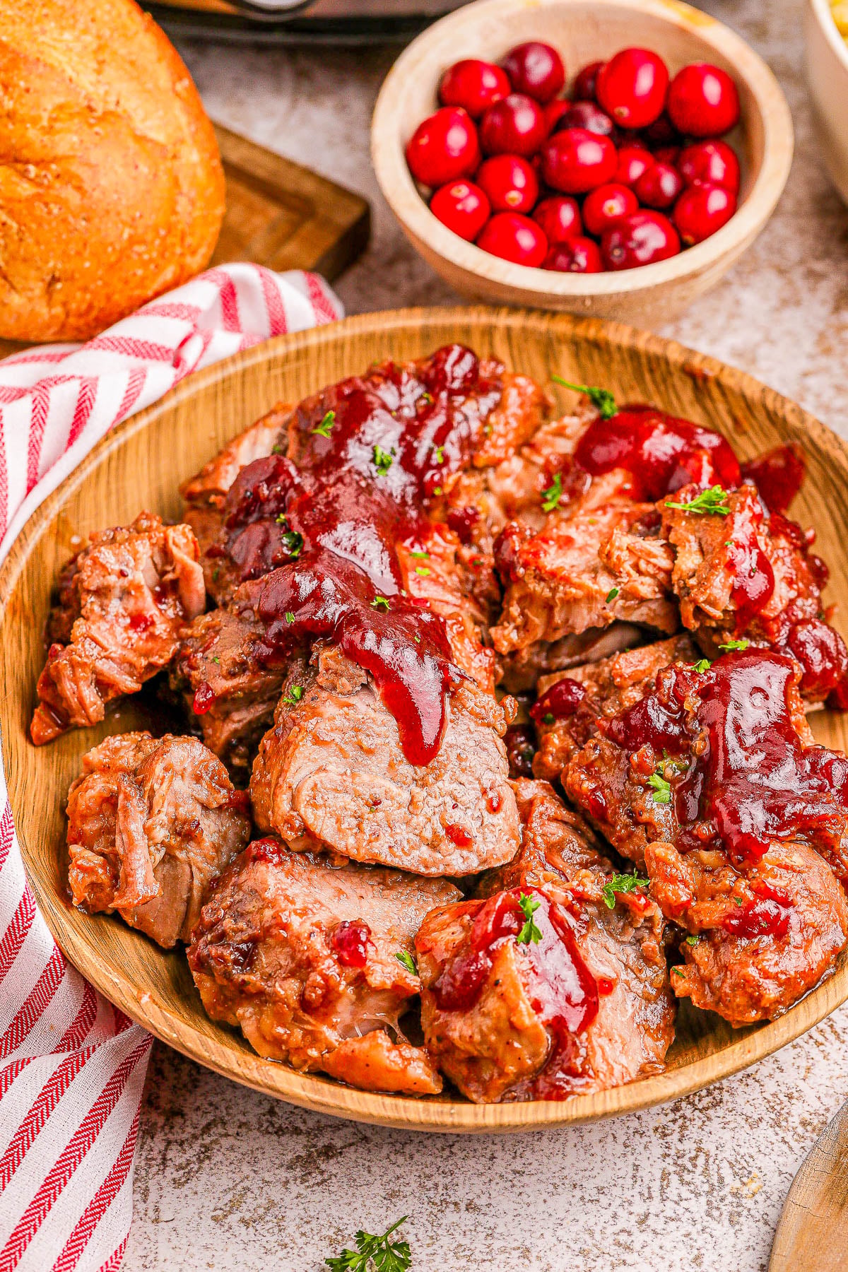Slow Cooker Cranberry Pork Tenderloin Sliced pork tenderloin covered with cranberry glaze in a wooden bowl with cranberries and bread behind