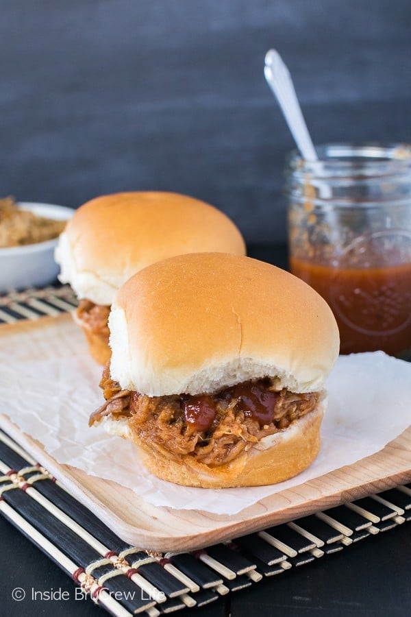 Easy Slow Cooker Pulled Pork Sandwiches Two finished pulled-pork sandwiches on soft rolls sit on a wooden board beside a jar of barbecue sauce