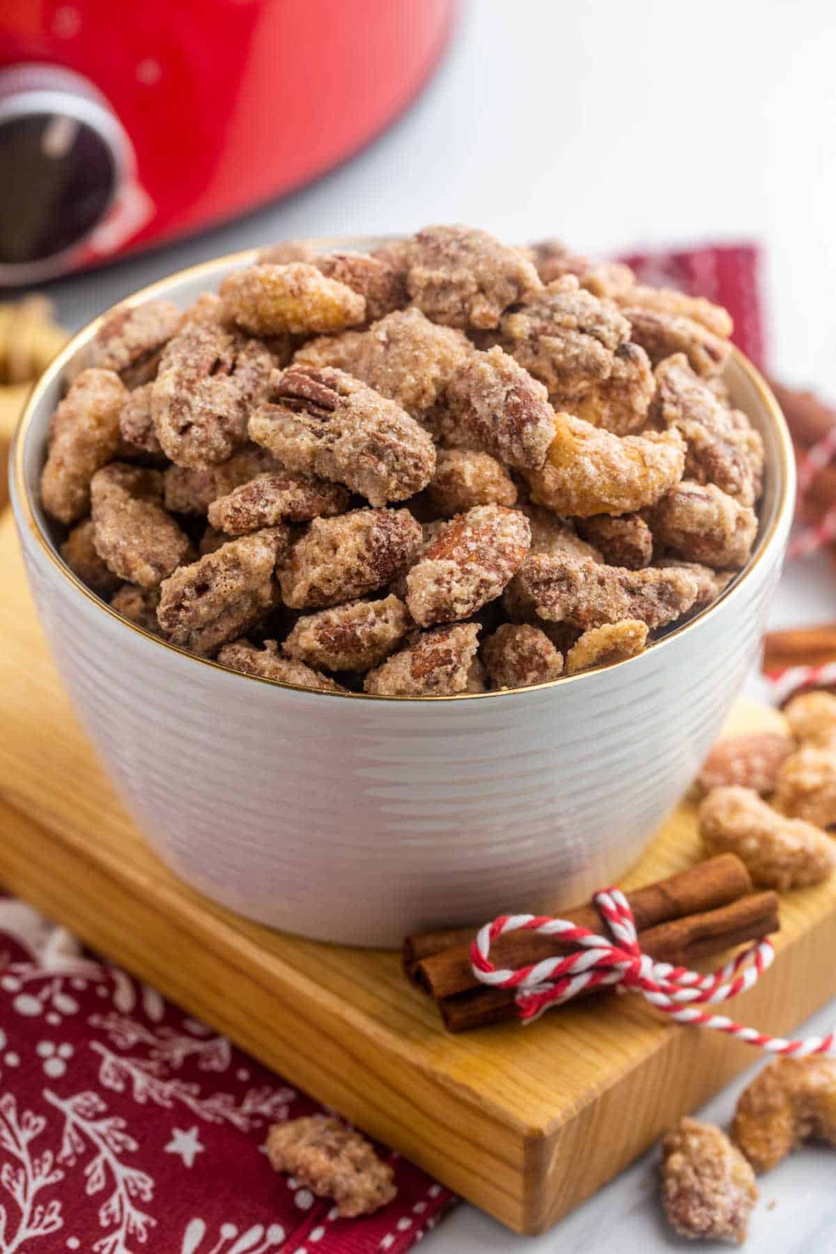Easy Crockpot Candied Nuts Recipe Cinnamon-sugar candied nuts in a bowl on red holiday textiles beside a slow cooker and cinnamon sticks