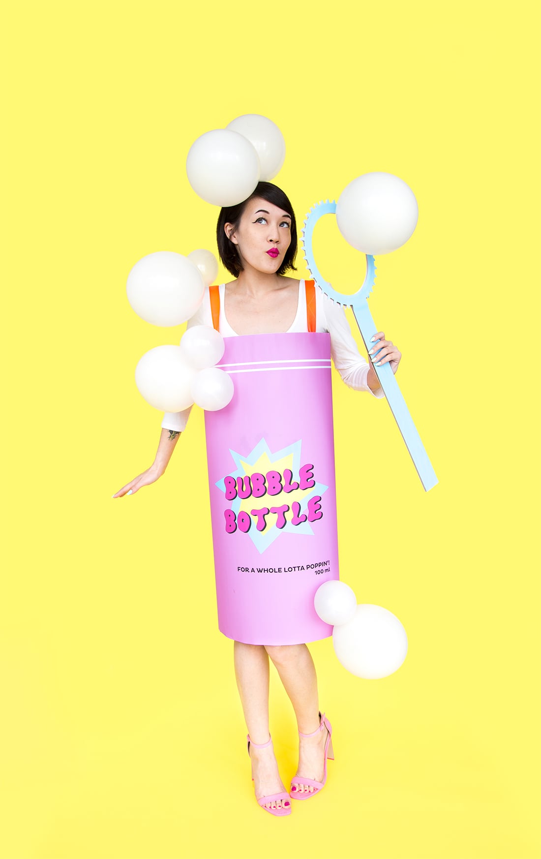 Adult wearing a pink bubble bottle costume with white balloon bubbles and holding an oversized blue bubble wand