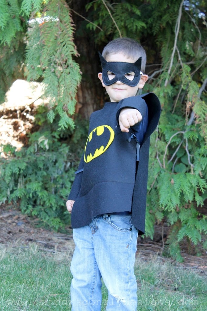 Child wearing a black felt Batman-style superhero tunic with yellow emblem, black mask and cape outdoors