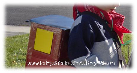 Child seen from behind wearing a brown cardboard train costume with a yellow side panel and source watermark
