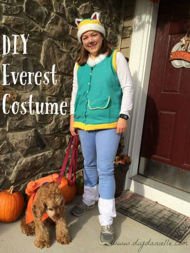 Adult wearing a teal Everest Paw Patrol vest, hat and leg warmers beside a costumed dog and pumpkins outdoors