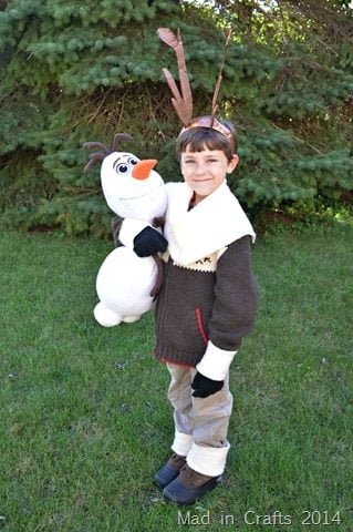 Child dressed as Sven with brown antler headband, shaggy cuffs, white collar and Olaf plush on green grass