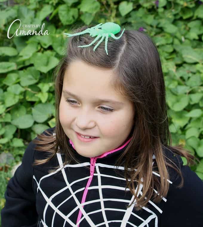 Child wearing a black hoodie marked with a white spiderweb and a green spider clipped into her brown hair