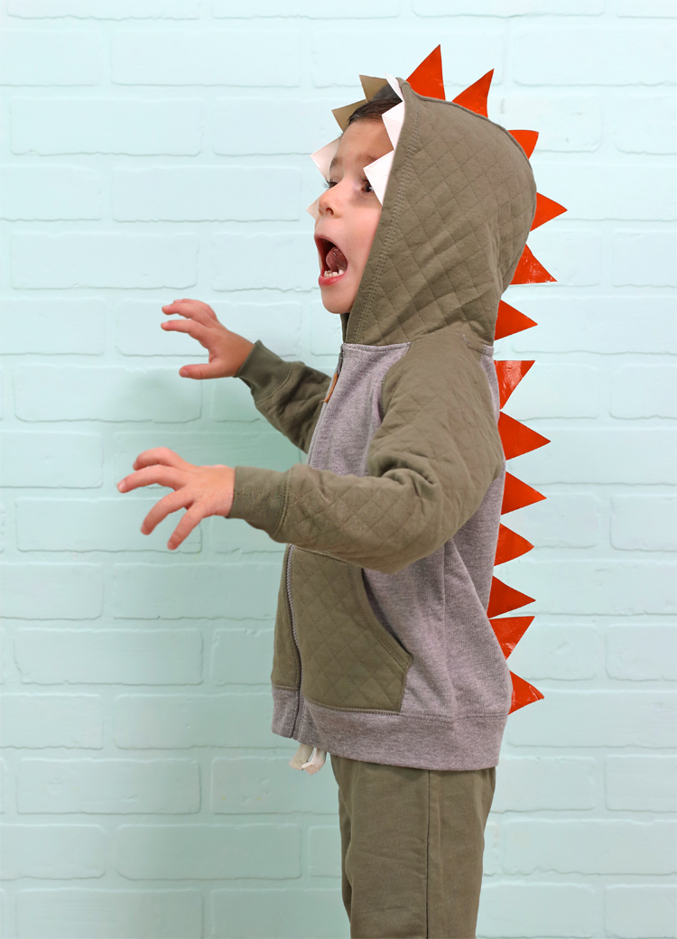 Child wearing a gray-green hooded dinosaur costume with white taped teeth and red-orange triangular spikes down the back