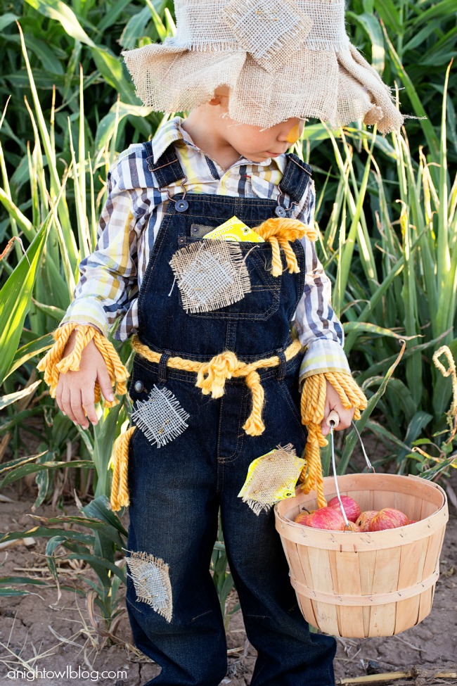 Child in patched denim overalls, plaid shirt and a floppy burlap scarecrow hat holding a wooden apple basket