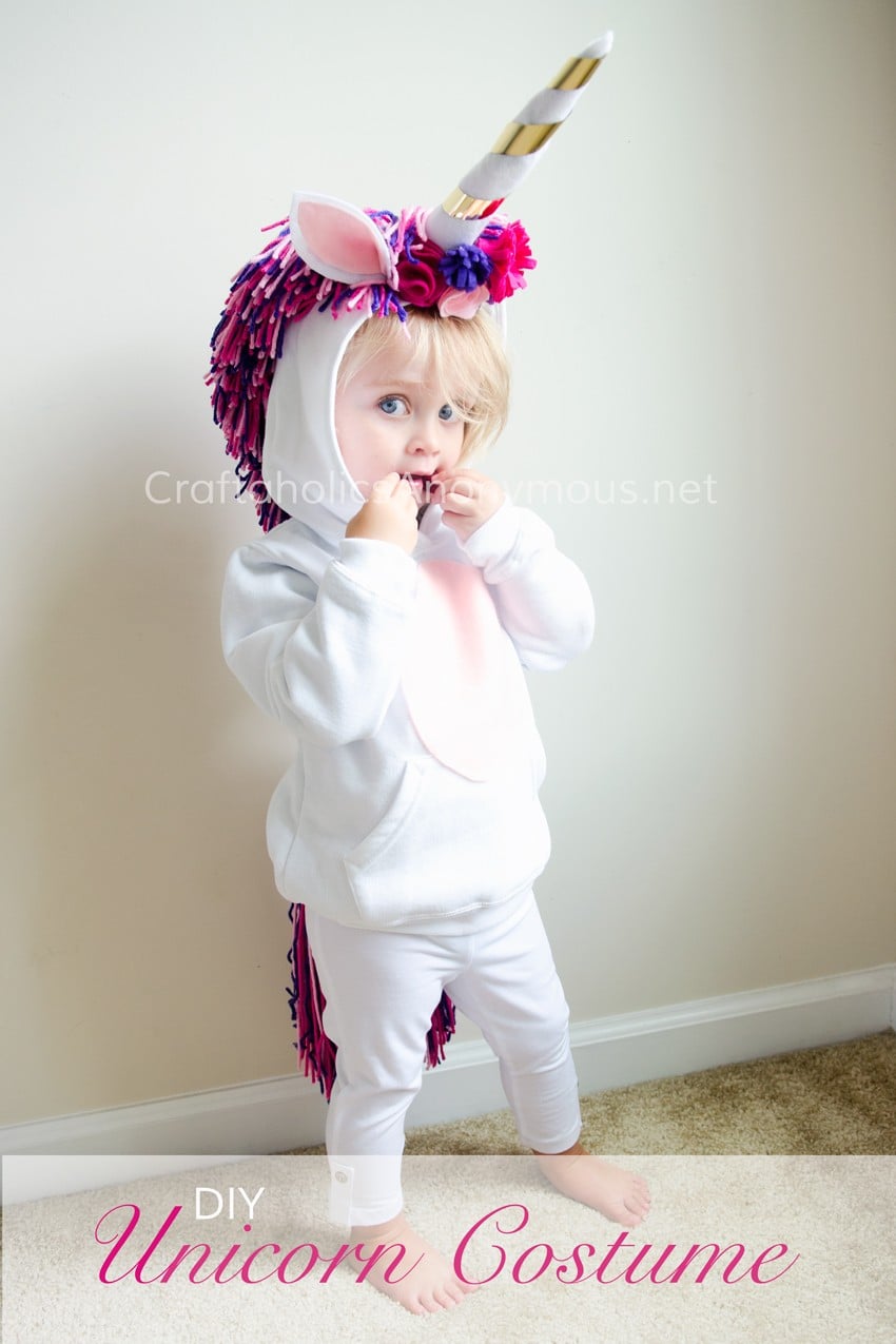 Child wearing a white unicorn hoodie costume with a gold-striped horn, felt ears, flowers and a pink-purple yarn mane