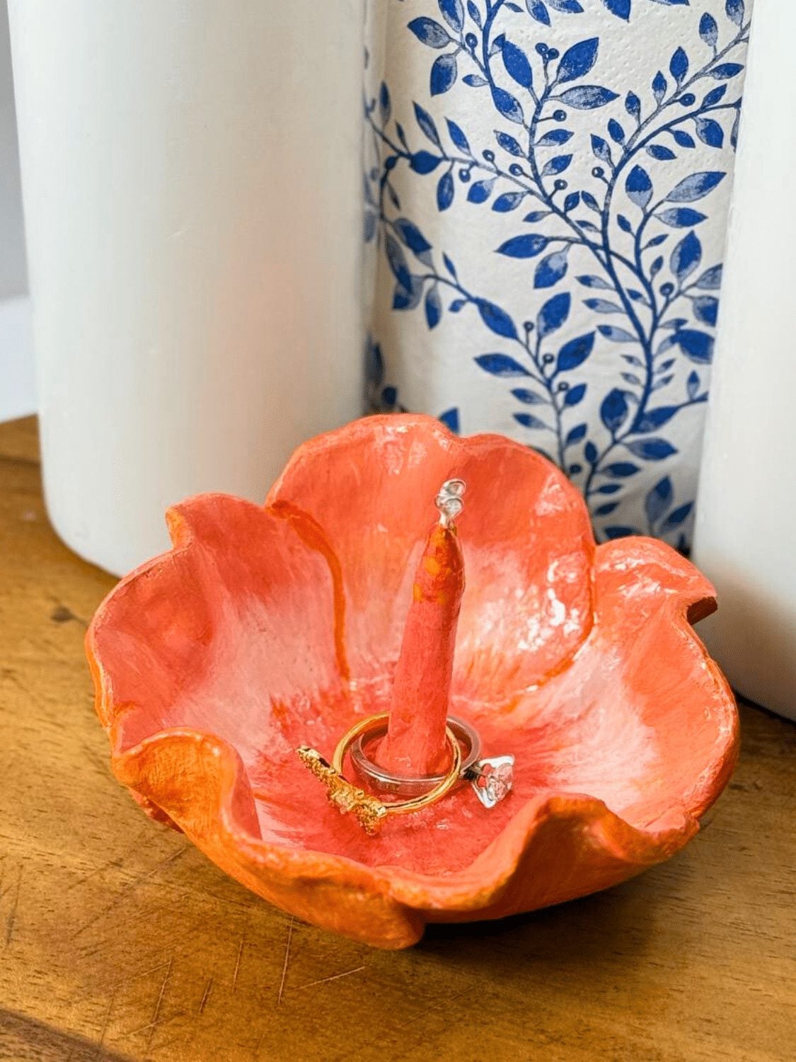 Coral orange air dry clay flower ring holder with a ring resting on the center post, beside a blue and white vase