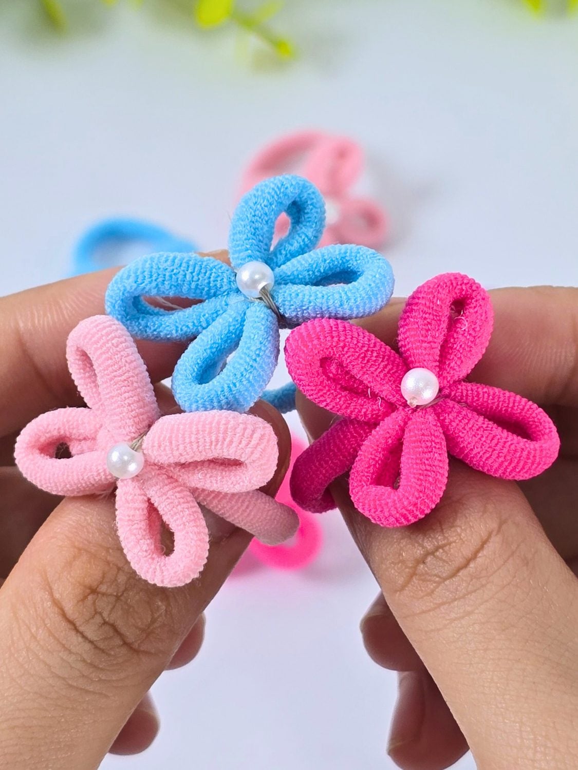 Pink and blue hair tie flower rings with pearl centers held between two fingertips against a white background