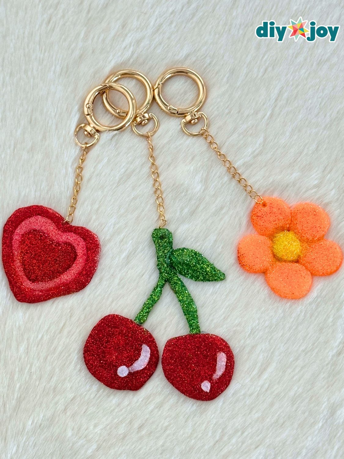 Three glitter hot glue keychains shaped like red cherries, a red heart and an orange daisy on gold chains, on white fur