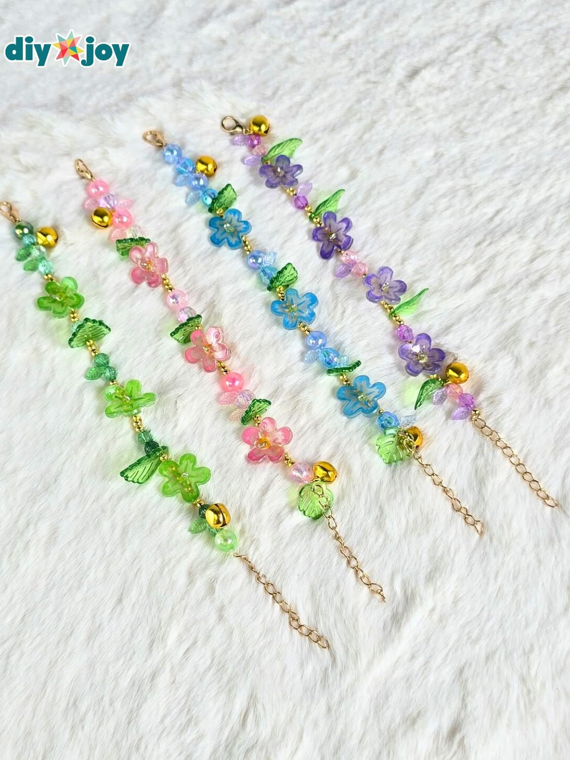 Four dainty shrink plastic flower bracelets in pink, blue, purple and green on gold chains laid in a row on white fur