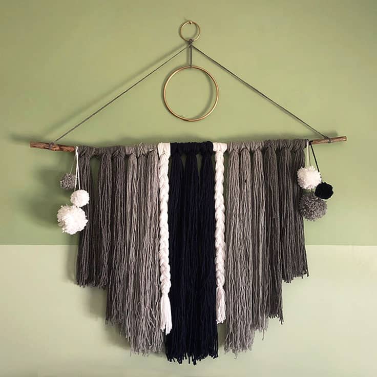 Bedroom wall art idea, a boho yarn wall hanging with gray and black fringe, braids and pom poms on a branch