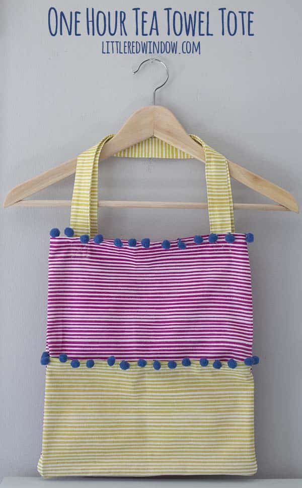 Tea towel tote bag in magenta and lime stripes with blue pom-pom trim and striped handles, hanging on a wooden clothes hanger