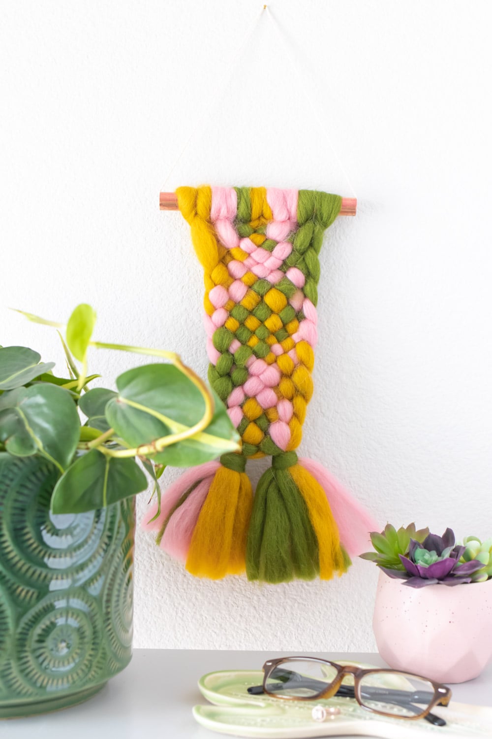 Small woven wall hanging of chunky pink, mustard and green roving on a copper pipe above a plant, succulents and glasses