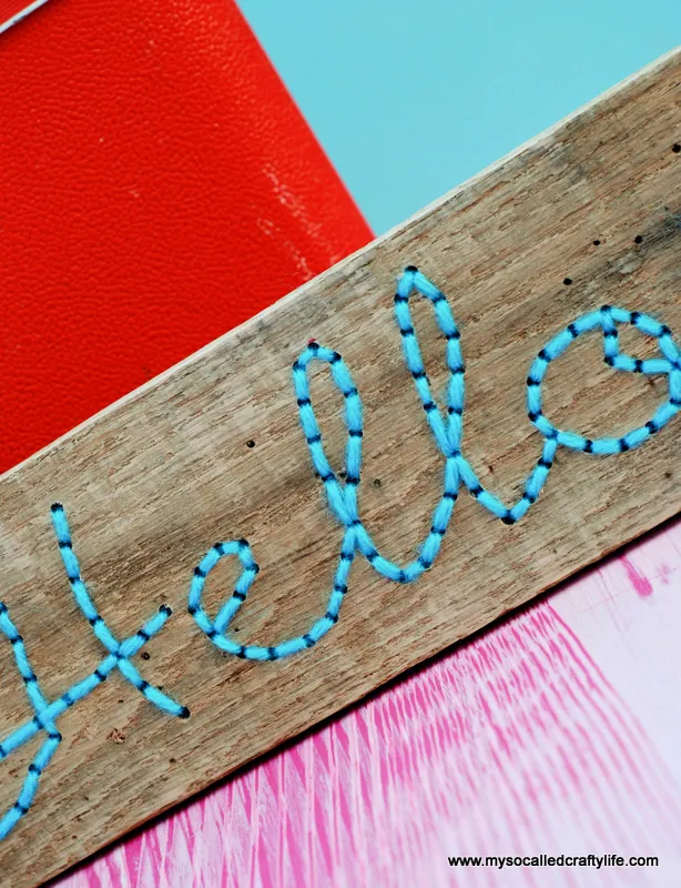 DIY embroidered pallet wood art, the word hello back stitched in aqua yarn on a narrow board beside a vintage suitcase