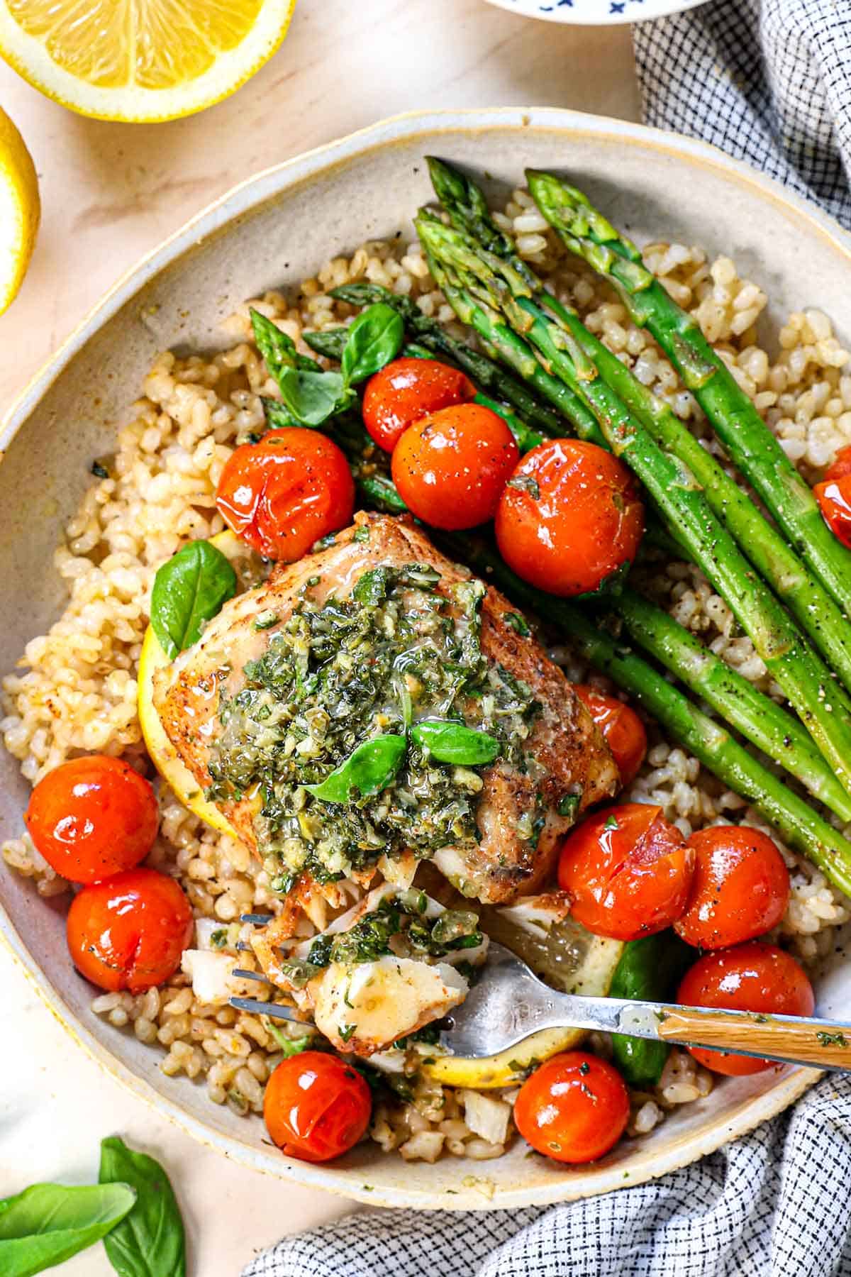 Baked cod fillet topped with herb butter beside roasted cherry tomatoes, asparagus and grains