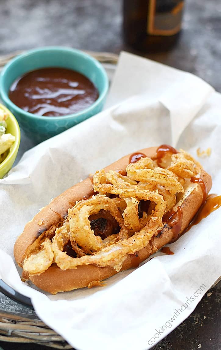Bacon-wrapped barbecue hot dog in a bun topped with crispy onion rings and BBQ sauce in a basket