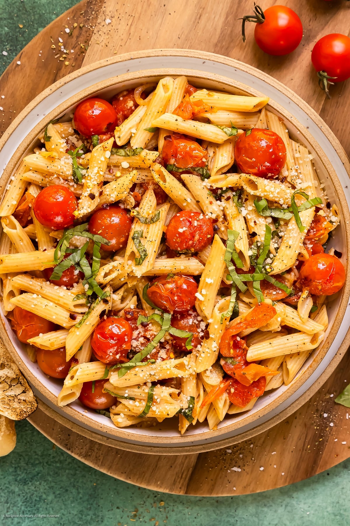 Bowl of penne with blistered cherry tomatoes, sliced basil and grated Parmesan on a wooden board