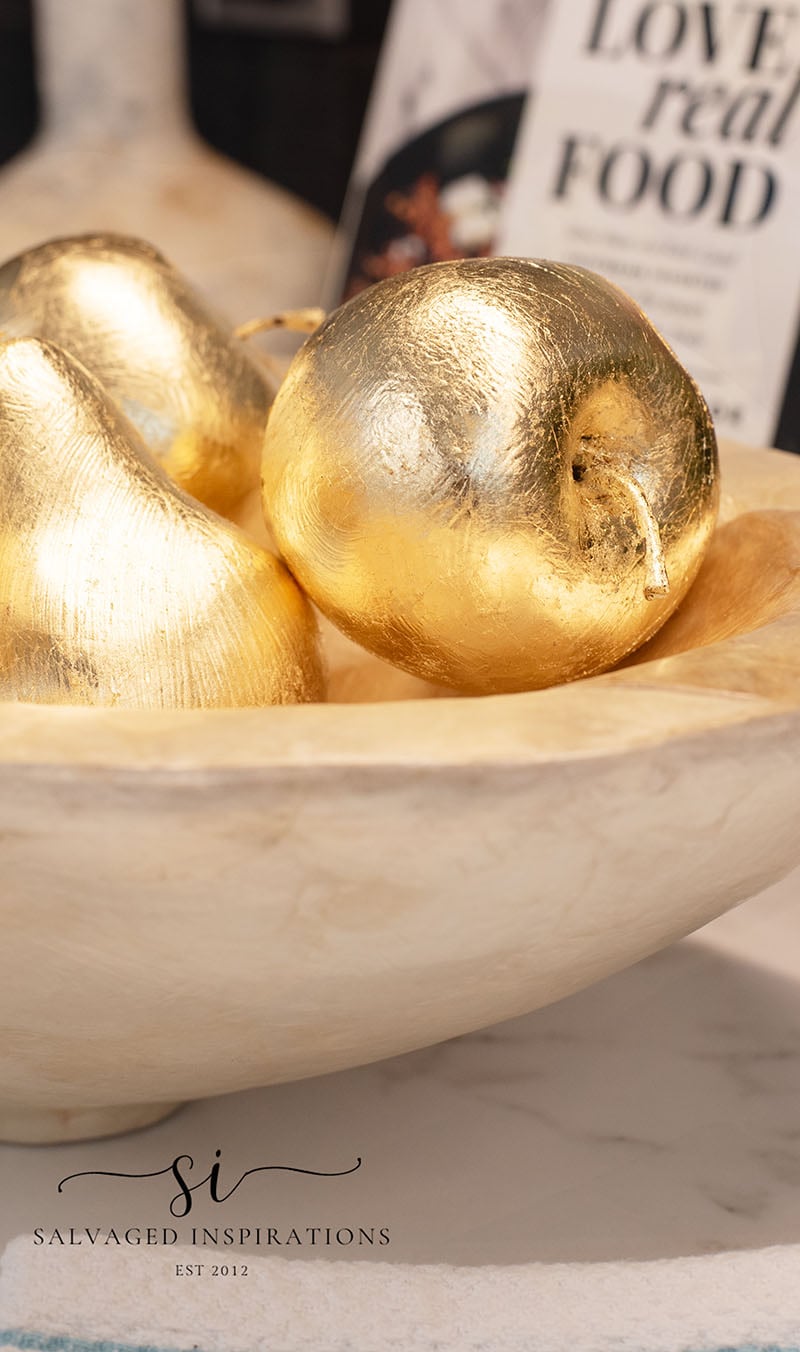 DIY gilded apples, a plastic apple being covered in shiny gold leaf sheets by a gloved hand on brown paper with gold flakes
