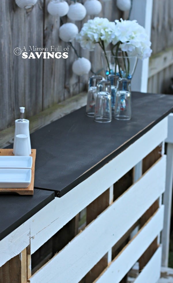 DIY Patio Bar | Pallet Bar with Chalkboard Top White paper lantern string lights glowing at night over a dark patio bar counter with glasses by a fence