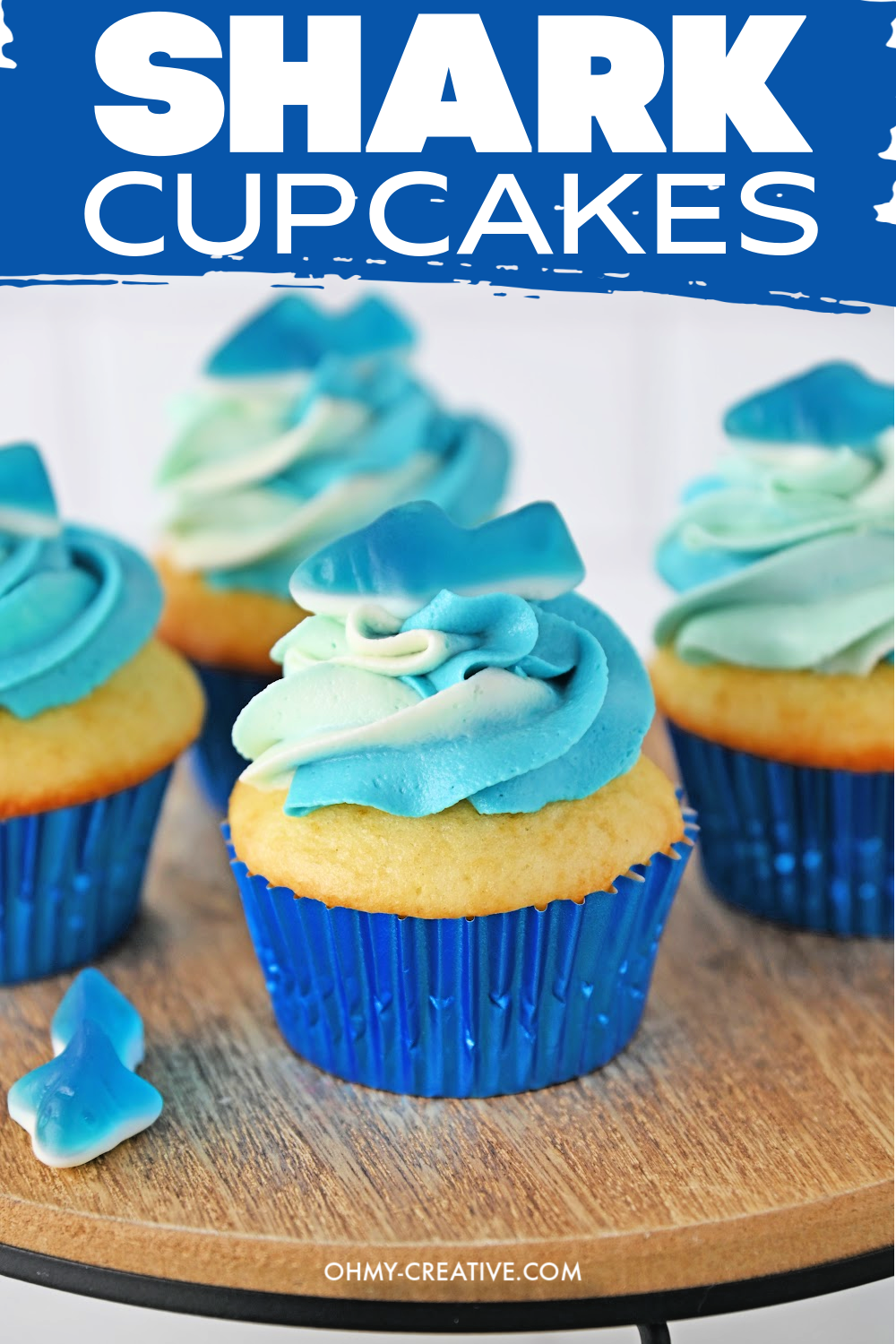 Cupcakes in blue foil liners with white, teal and blue ombre swirls, each topped with a blue gummy shark