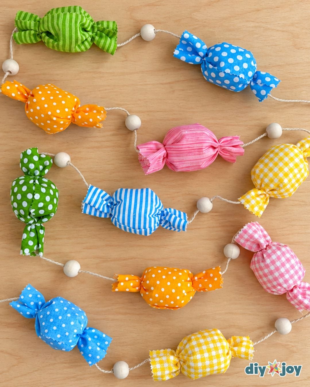 DIY candy garland of polka dot, striped and gingham fabric candies strung on twine with wooden beads across a wood table