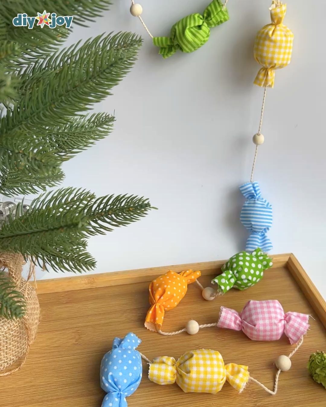 DIY candy garland hanging down a white wall onto a wooden tray, next to a green pine branch in a burlap pot