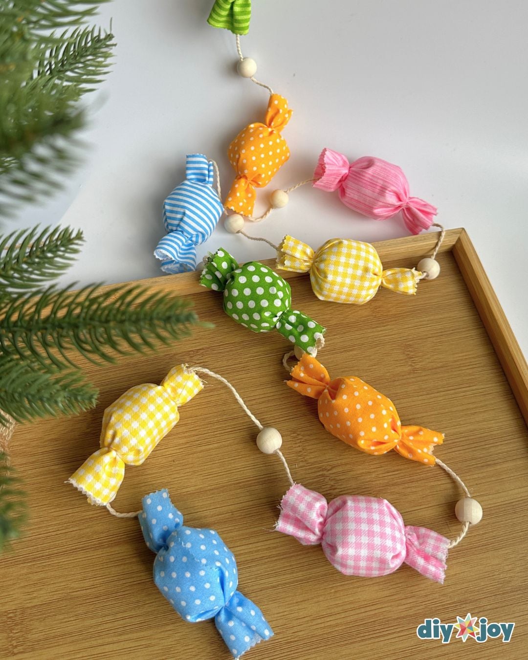 Fabric candy garland with wooden beads curling across a wooden tray beside green pine branches for holiday decor