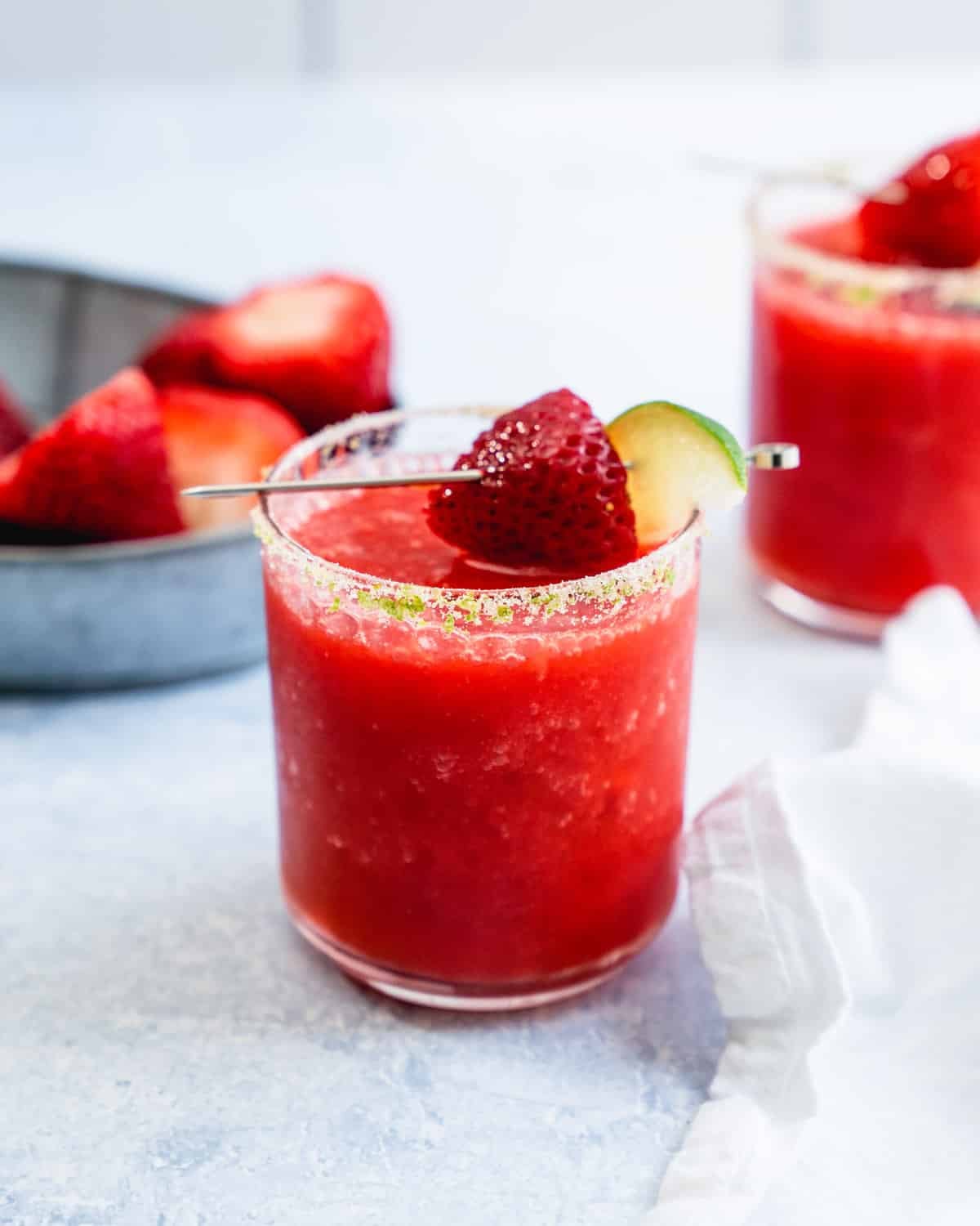 Blended frozen strawberry margarita with a lime salt rim, strawberry and lime garnish beside fresh berries