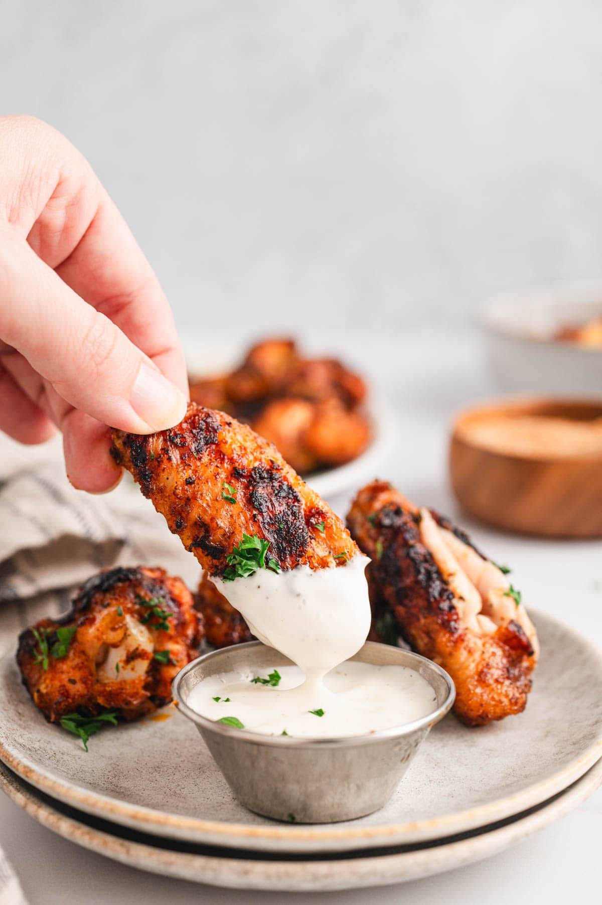 Grilled Chicken Wings Grilled chicken wing with charred spice rub being dipped into a small metal cup of ranch, more wings on a plate behind it
