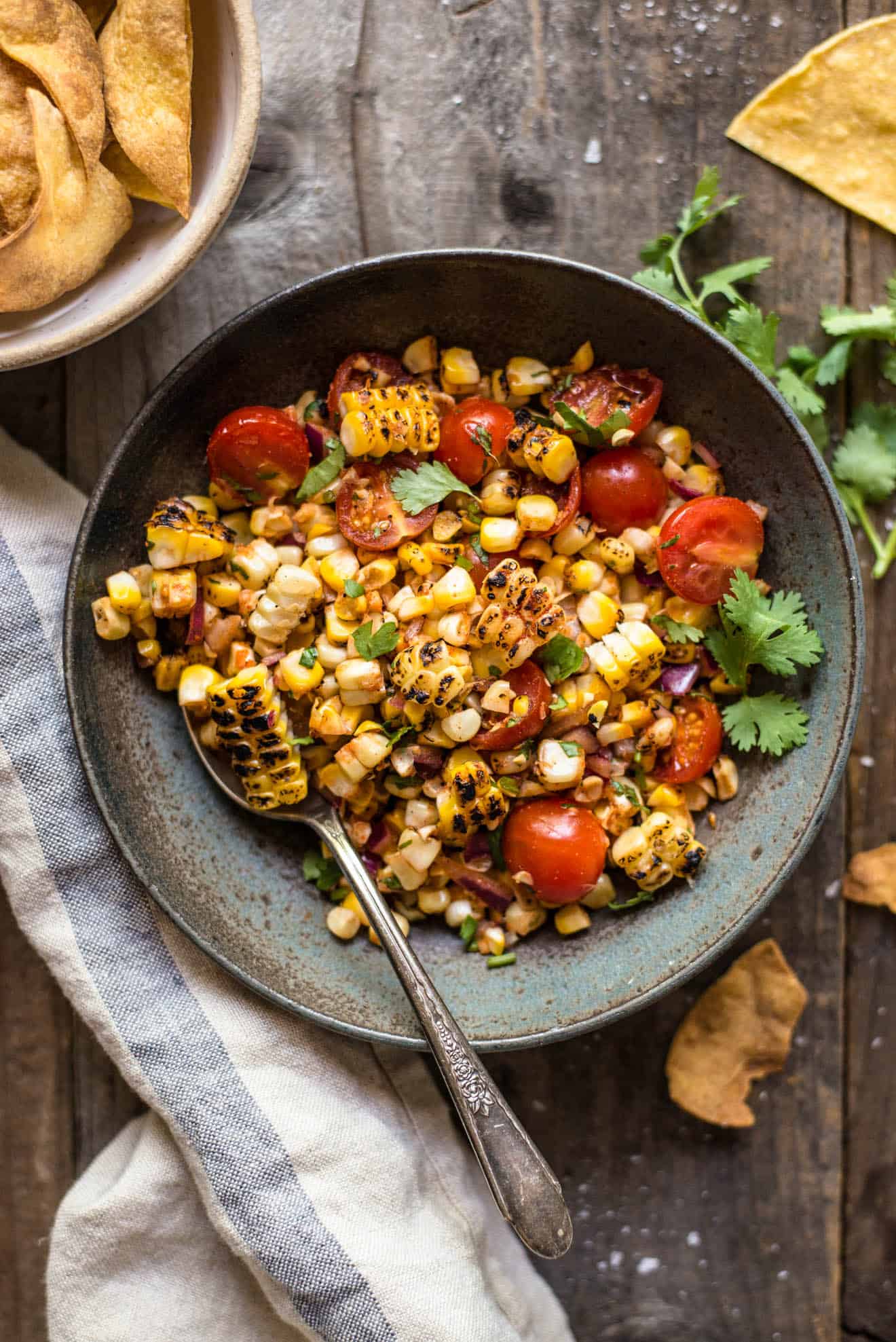 Grilled corn salsa with charred kernels, cherry tomatoes, red onion and cilantro in a dark bowl with tortilla chips