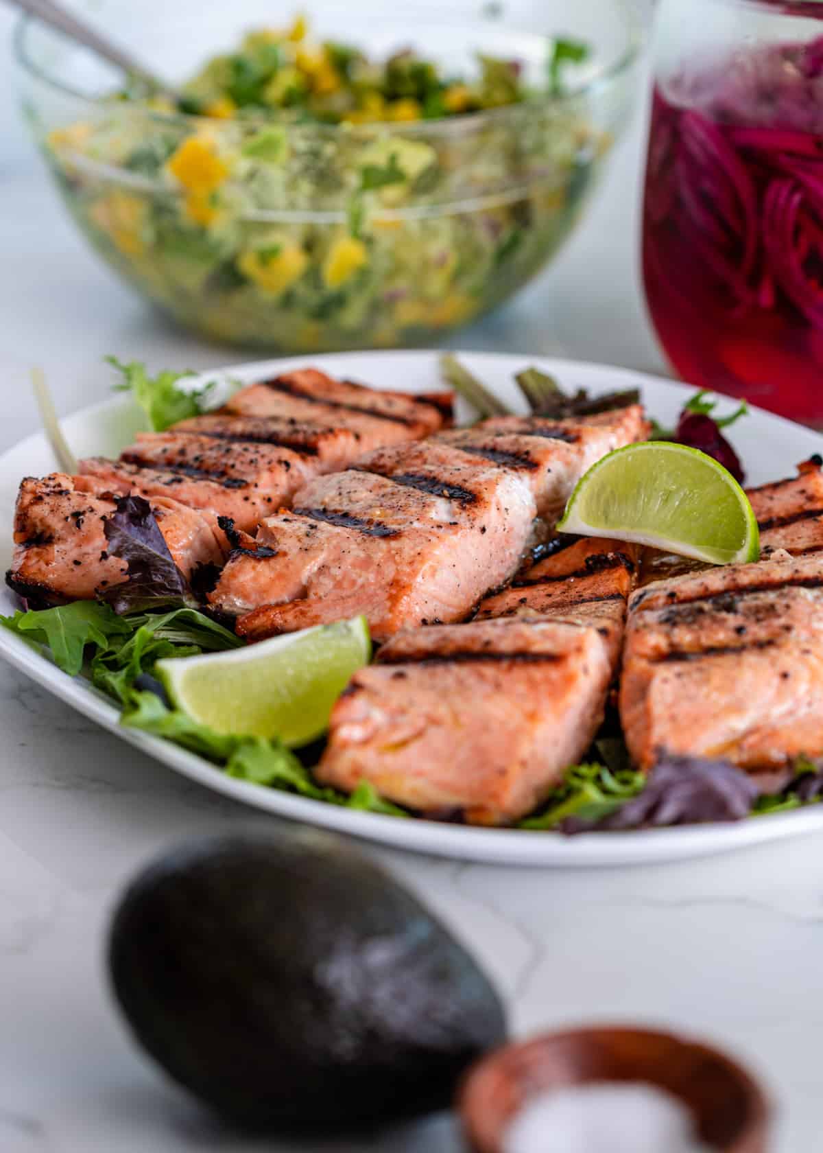 Grilled Mango Salsa Salmon Grill-marked salmon pieces on a bed of greens with lime wedges, a glass bowl of mango avocado salsa behind them