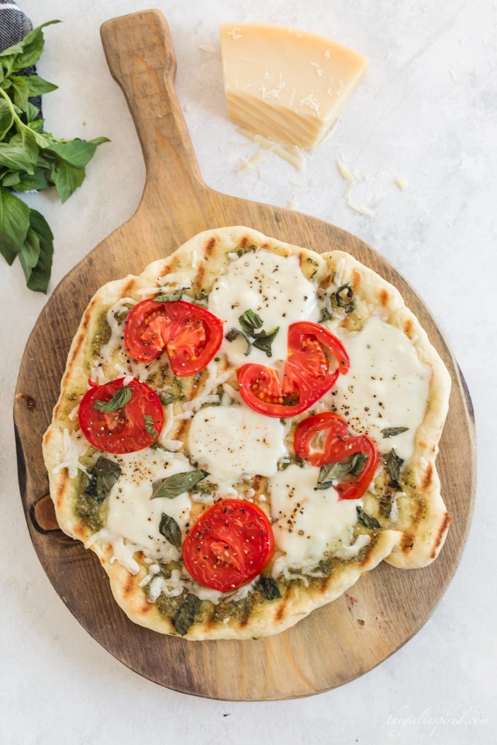 Grilled margherita pizza with melted mozzarella, tomato slices, basil and pesto on a round wooden board with a wedge of parmesan