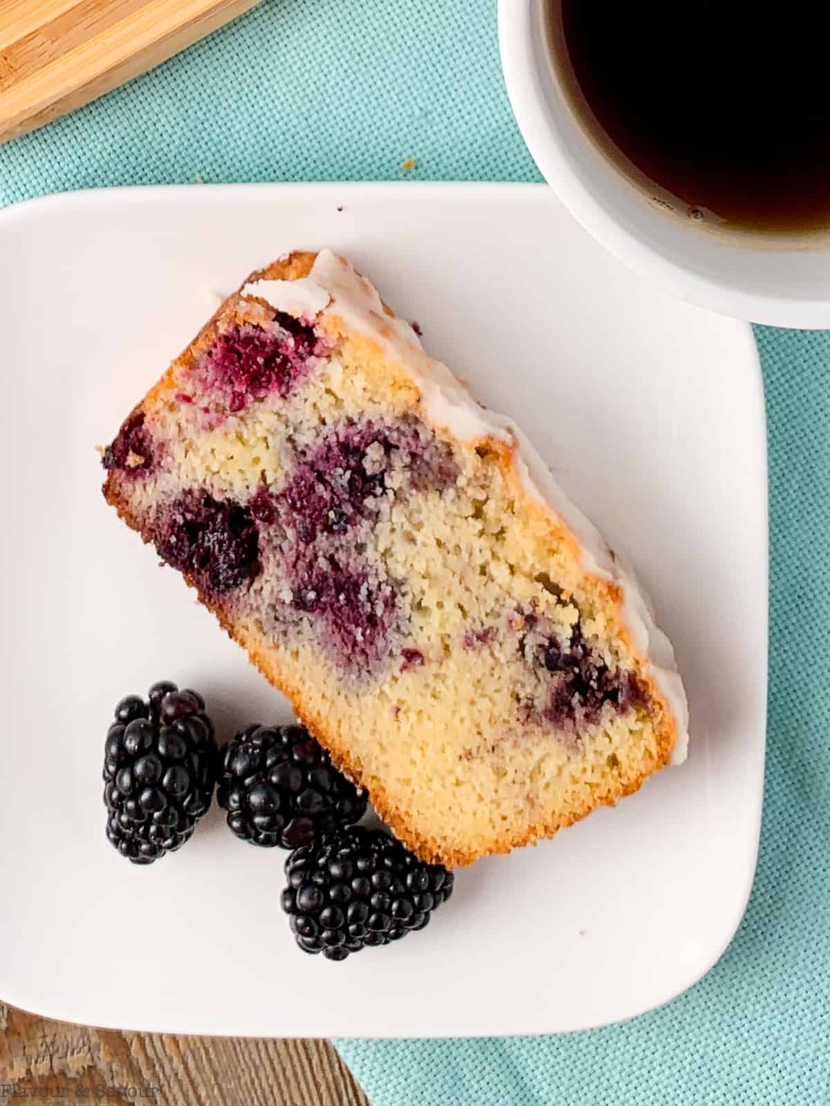 Slice of keto blackberry bread with white lemon glaze on a plate with fresh blackberries and coffee