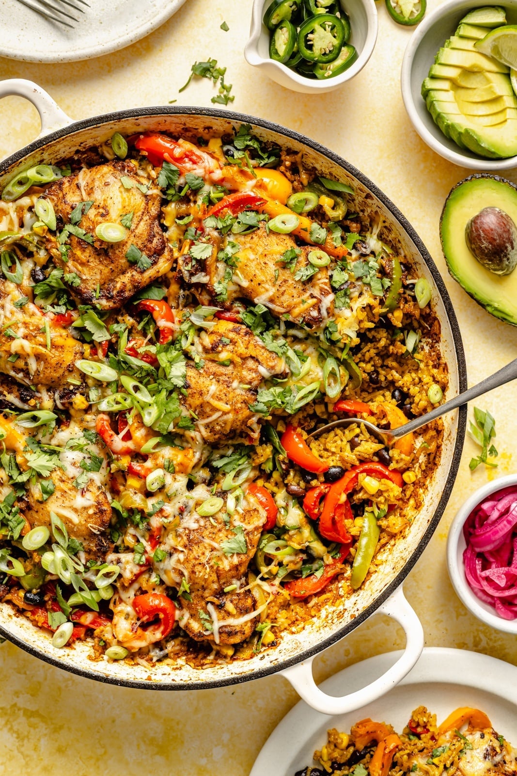 Large skillet of fajita chicken and rice with peppers and green onion, sides of avocado and jalapeno
