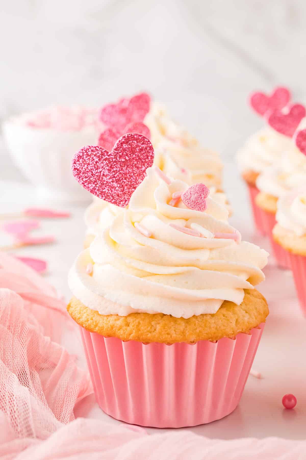 Cupcake in a pink liner with a white frosting swirl, pink sprinkles and pink glitter heart toppers