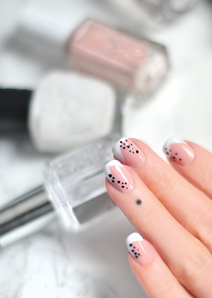 Abstract French Tip Nails with Dots | Quick Nail Art Idea Abstract French tip manicure with angled white tips and clusters of tiny black dots on pink nude nails, beside polish bottles