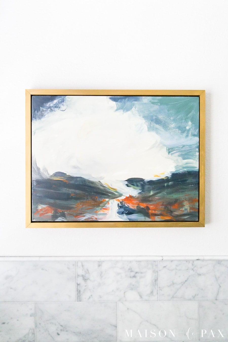 Abstract landscape painting in blue, white, navy and orange in a thin gold frame above marble bathroom tile