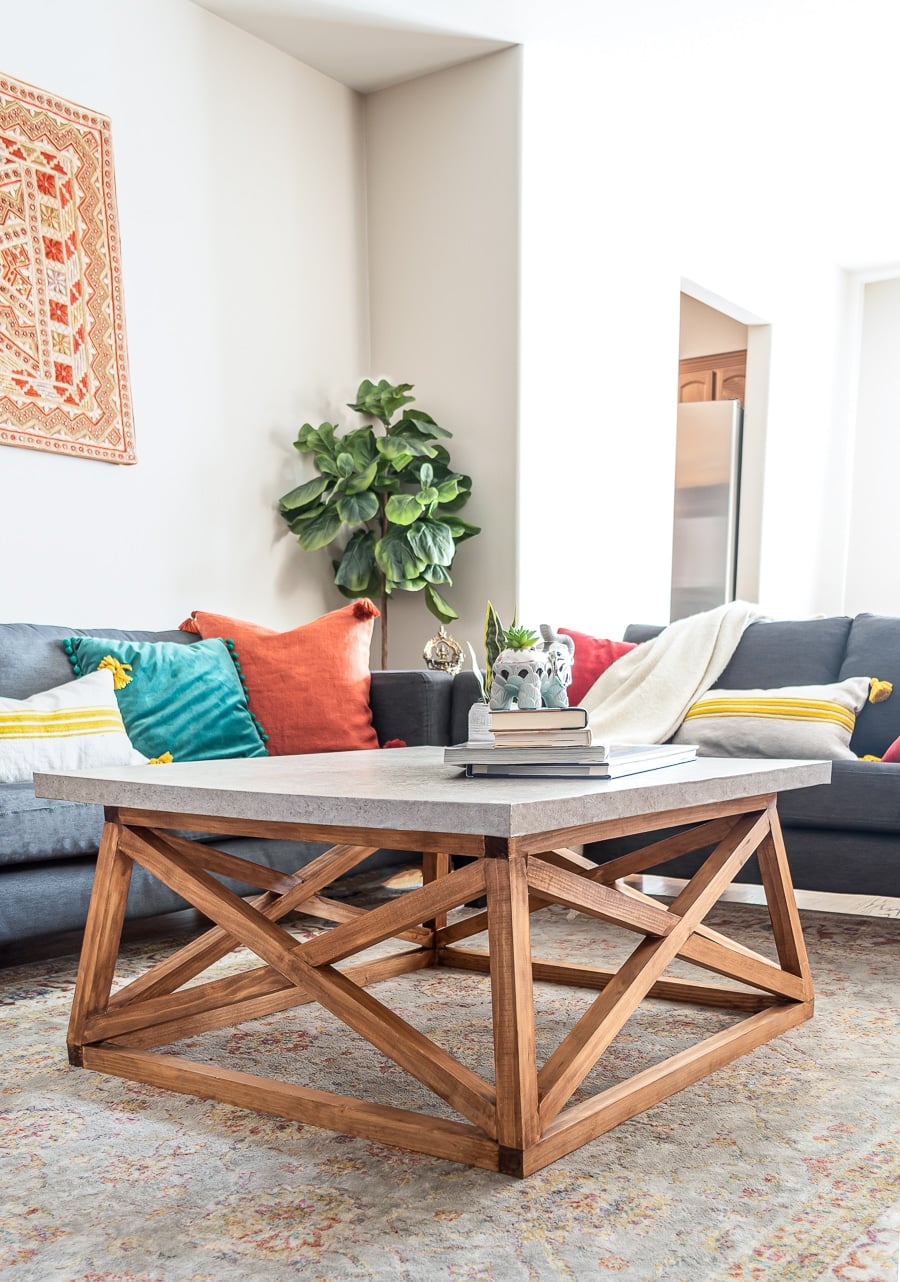 Square coffee table with a gray faux concrete top on an angled wood X base with books and a plant in a bright family room