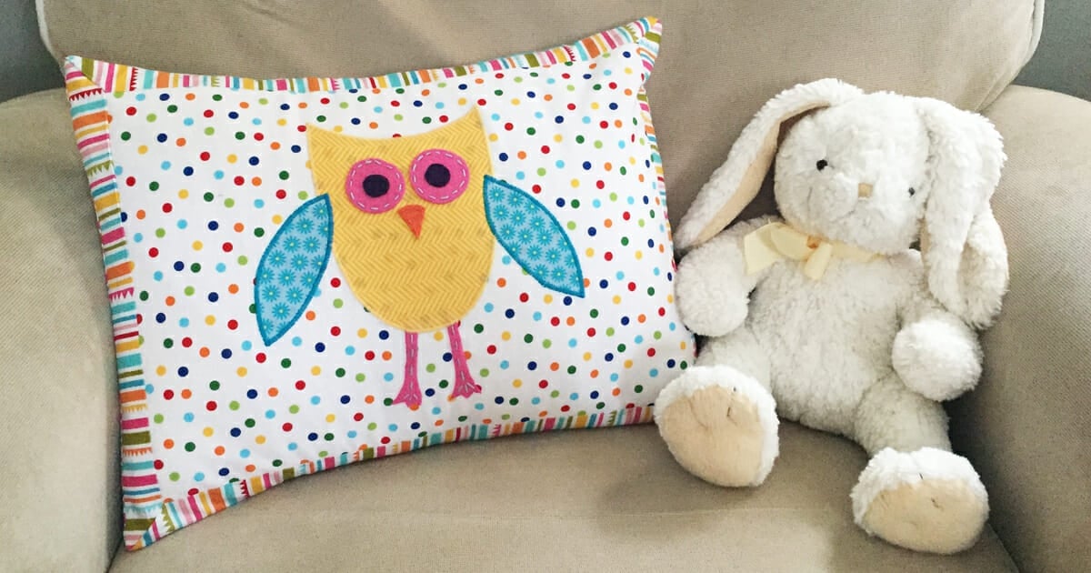 Rectangle nursery pillow with a yellow owl applique on rainbow polka dot fabric and a striped border, by a white bunny toy