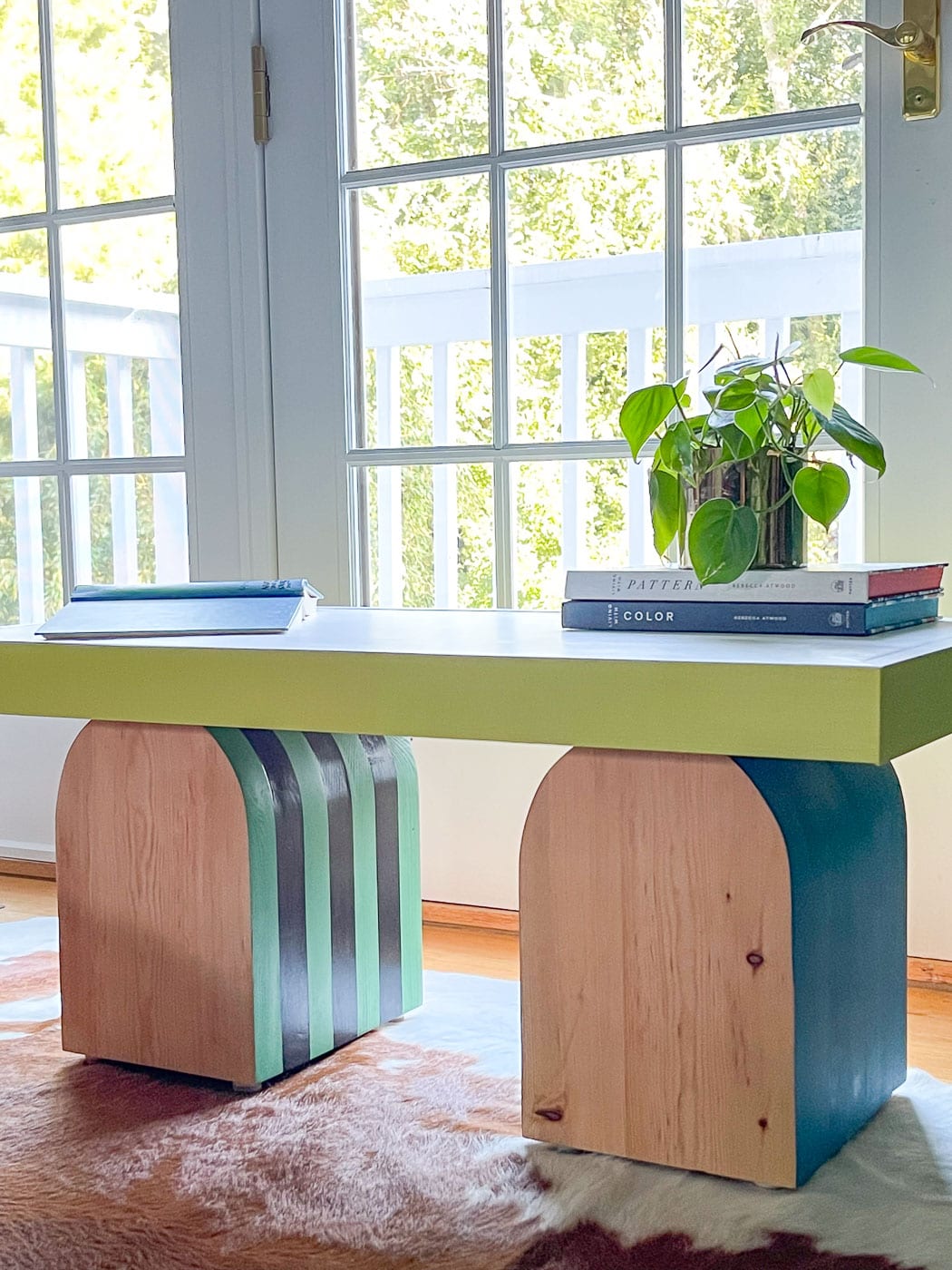 Coffee table with a lime green top on two chunky arched wood legs painted with teal and green stripes by a sunny window
