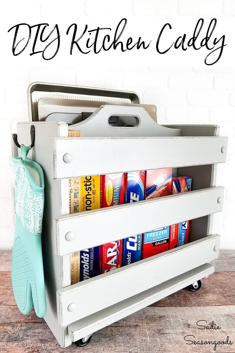 Gray wooden caddy on casters holding baking sheets on top and boxes of foil and wrap below, with oven mitts on side hooks
