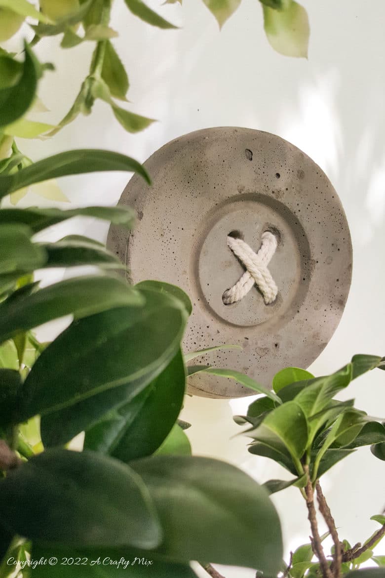 Big Concrete Button Hooks | Craft Room Wall Hooks Round gray concrete button with four holes and a white cord stitched through in an X, hanging among green leaves