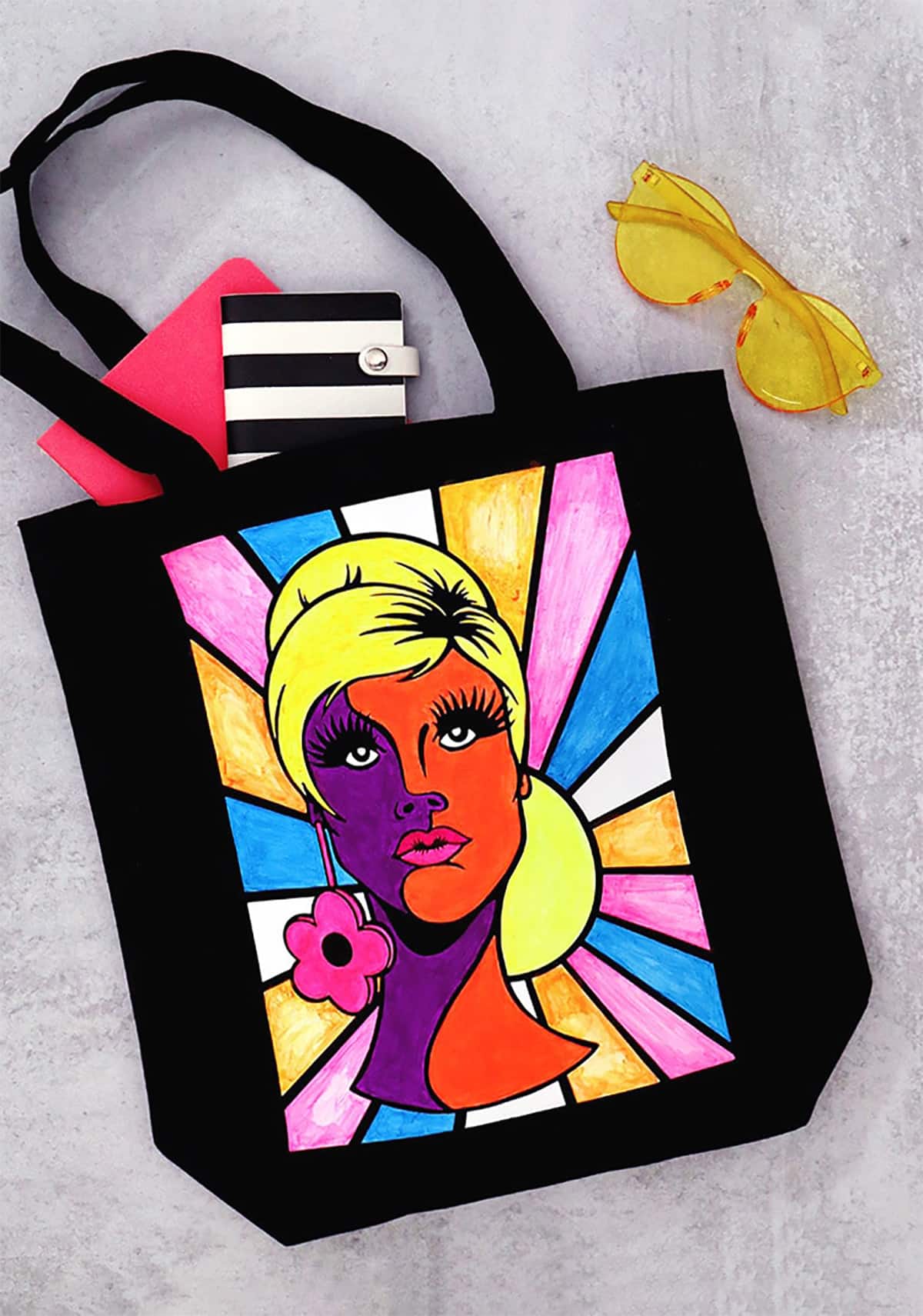 Black canvas tote with a painted retro lady face in neon pink, orange, purple and yellow next to yellow sunglasses