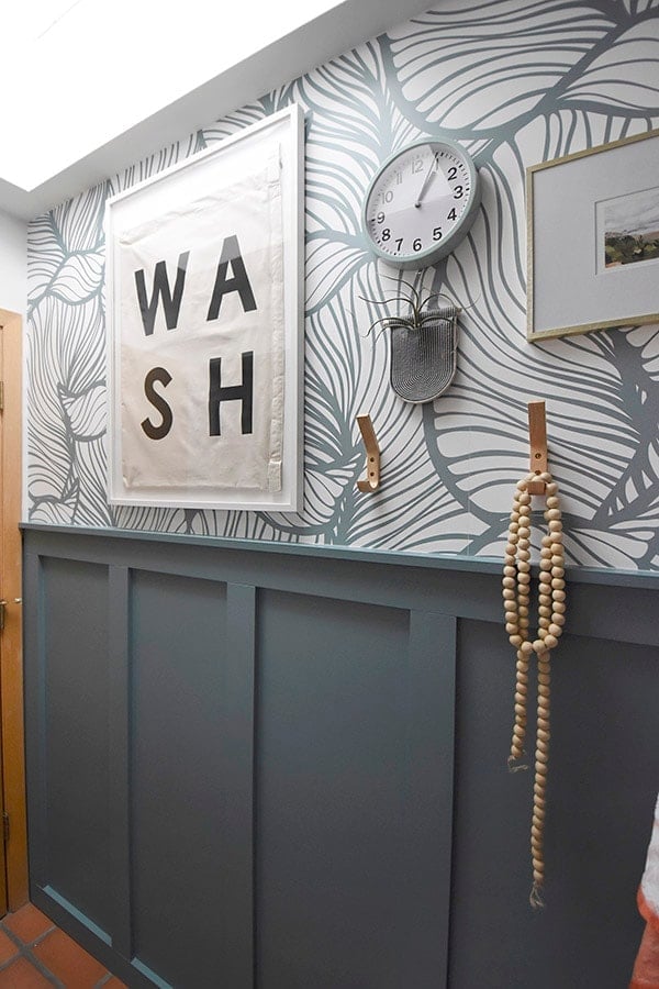 Bathroom wall with tall dark teal board and batten under gray leaf wallpaper, a framed WASH sign, clock and wood bead garland