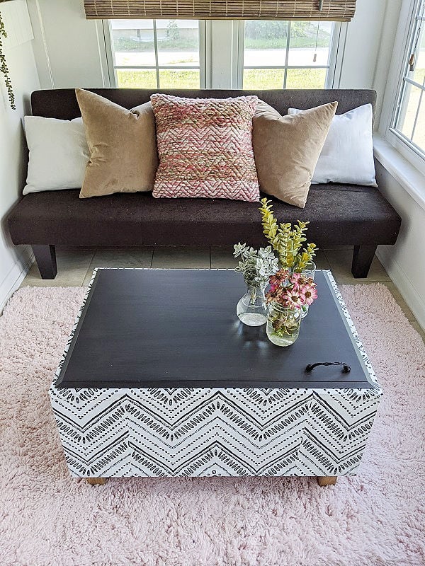 Coffee table storage bench covered in black and white zigzag wallpaper with a black top and flowers in a bright sunroom