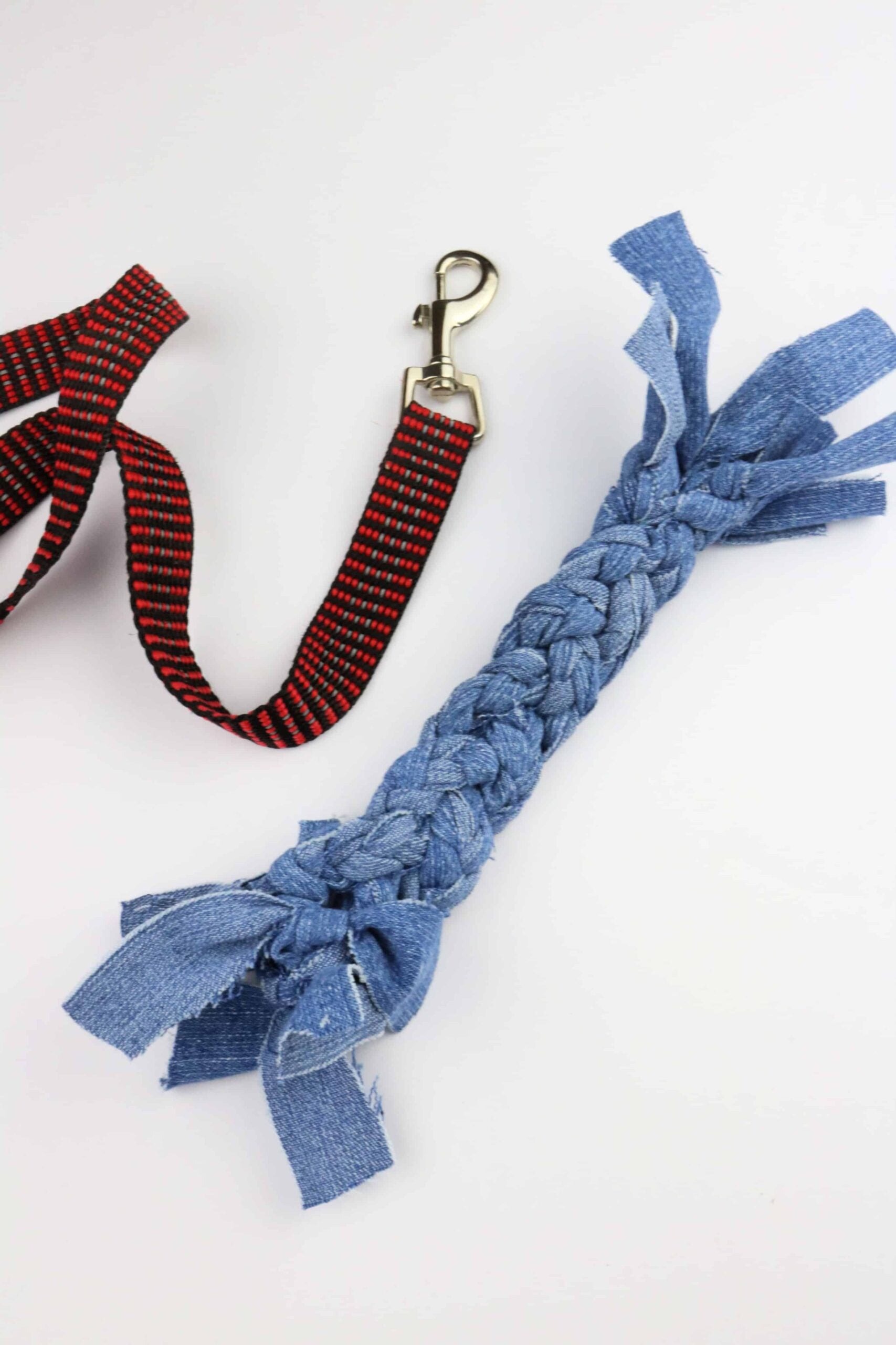 Braided light blue denim rope dog toy with knotted frayed ends lying beside a red and black dog leash on a white surface